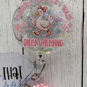 May include: A round, clear badge reel with a cartoon duck wearing a pink bow and boots. The text "SORRY I'M LATE I WAS" and "DILLY DALLYING" are in pink. The reel has a retractable cord and a pink, sparkly charm.