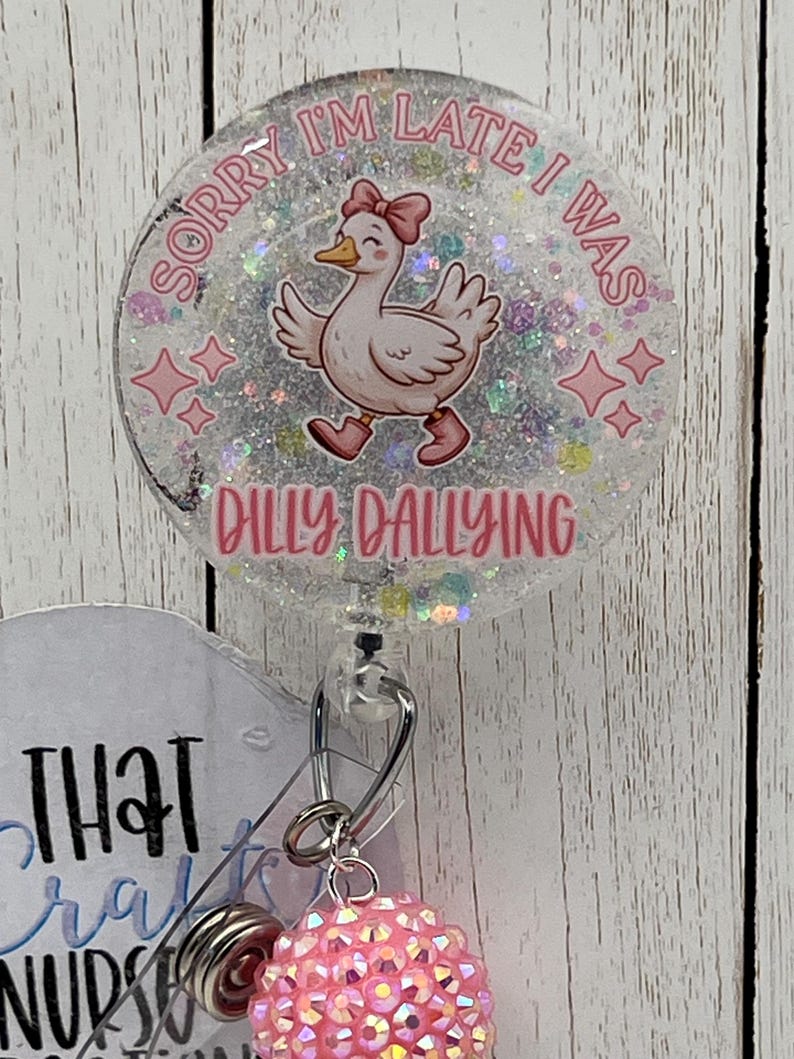 May include: A round, clear badge reel with a cartoon duck wearing a pink bow and boots. The text "SORRY I'M LATE I WAS" and "DILLY DALLYING" are in pink. The reel has a retractable cord and a pink, sparkly charm.