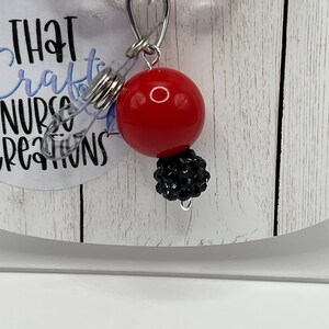 May include: A red, round badge reel with a black beaded accent. The reel has a clear plastic strap and a metal clip. The text "THAT crafts NURSE creations" is visible on the badge reel.