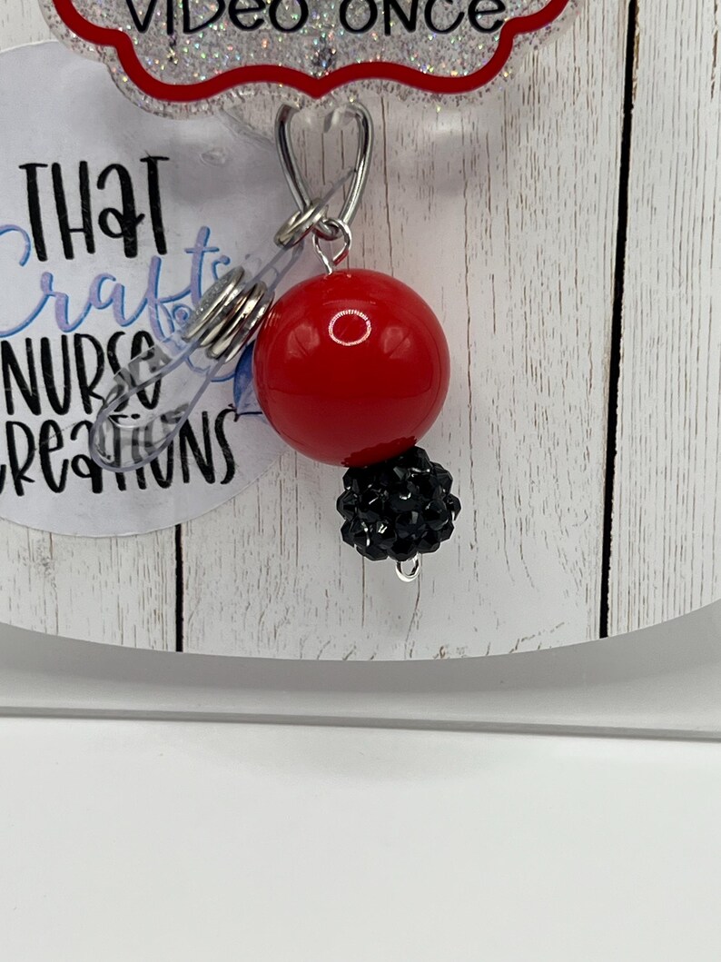 May include: A red, round badge reel with a black beaded accent. The reel has a clear plastic strap and a metal clip. The text "THAT crafts NURSE creations" is visible on the badge reel.