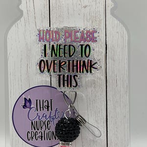 May include: A retractable badge reel with the text "HOLD PLEASE I NEED TO OVERTHINK THIS". The reel has a black and pink beaded charm and a purple circle with the text "THAT Crafty NURSE CREATION". The background is a white wooden plank design.