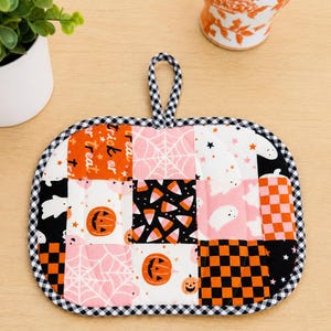 May include: A square quilted potholder featuring a patchwork design of Halloween-themed fabric squares in orange, black, white, and pink. Patterns include pumpkins, ghosts, spiderwebs, candy corn, and checkers, finished with a black and white gingham trim.