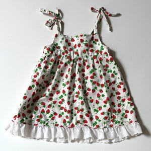 Könnte beinhalten: Ein weißes ärmelloses Kleid mit einem durchgehenden Muster aus kleinen roten Erdbeeren und grünen Blättern. Das Kleid hat Schulterträger zum Binden und einen weißen Saum mit Lochstickerei-Rüschen, was eine schlichte Silhouette ergibt.