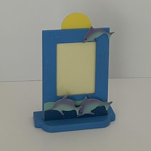 May include: A blue picture frame with a yellow sun and two dolphins. The frame has a rectangular opening for a photo and is decorated with two dolphins and a yellow sun. The frame is made of wood and is painted blue.