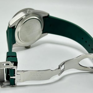 May include: A silver-toned watch with a green rubber strap. The watch band is open, revealing the clasp and the text "TUDOR GENEVE SWISS MADE STAINLESS STEEL". The watch is angled, showing the back of the watch face.