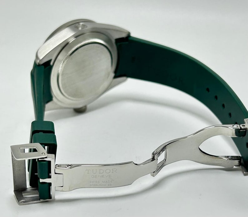May include: A silver-toned watch with a green rubber strap. The watch band is open, revealing the clasp and the text "TUDOR GENEVE SWISS MADE STAINLESS STEEL". The watch is angled, showing the back of the watch face.