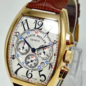May include: A gold-tone tonneau-shaped chronograph wristwatch with a white textured dial, black Arabic numerals, and three sub-dials. The watch features the text Franck Muller Geneve and Swiss Made, with blue and red accents, paired with a brown leather strap.