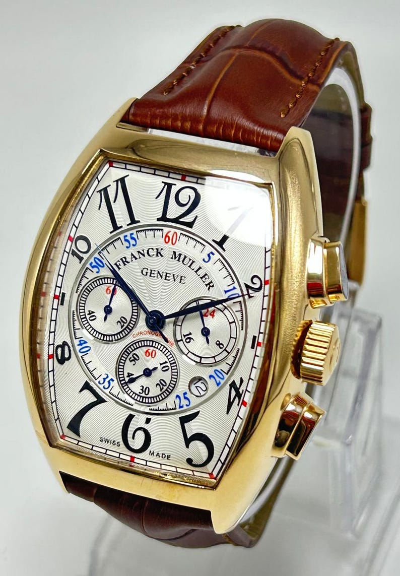 May include: A gold-tone tonneau-shaped chronograph wristwatch with a white textured dial, black Arabic numerals, and three sub-dials. The watch features the text Franck Muller Geneve and Swiss Made, with blue and red accents, paired with a brown leather strap.