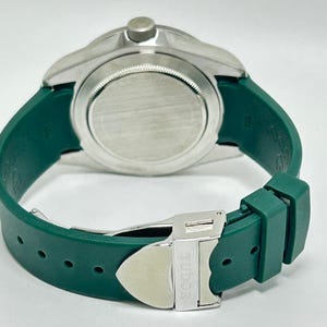 May include: A close-up of a silver-toned watch with a green rubber strap. The watch has a round face and a silver clasp with the word "TUDOR" engraved on it. The green strap is wrapped around the watch.