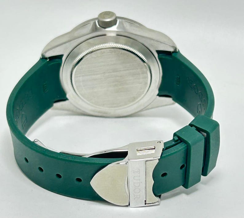 May include: A close-up of a silver-toned watch with a green rubber strap. The watch has a round face and a silver clasp with the word "TUDOR" engraved on it. The green strap is wrapped around the watch.