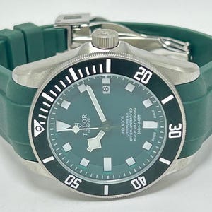 May include: A stainless steel Tudor Pelagos watch with a green dial and bezel. The watch has a green rubber strap and a date window at the 3 o'clock position. The watch is water resistant to 500m.