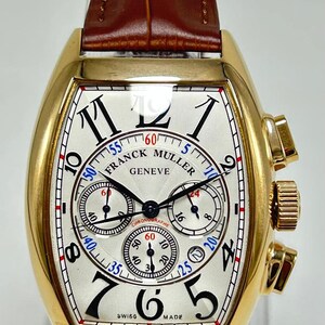 May include: A gold-toned tonneau-shaped chronograph wristwatch featuring a white textured dial with black Arabic numerals, three sub-dials, and the text Franck Muller Geneve. The watch includes a brown leather strap with a crocodile-like pattern.