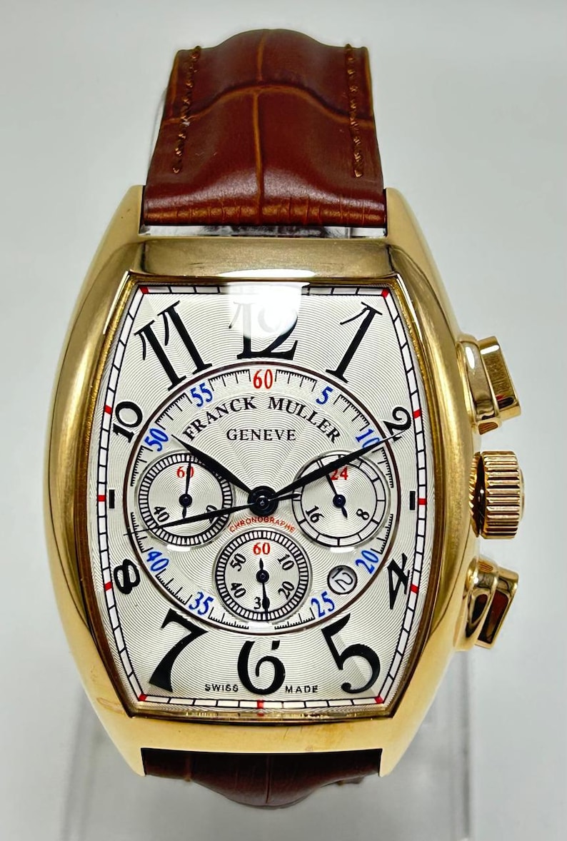 May include: A gold-toned tonneau-shaped chronograph wristwatch featuring a white textured dial with black Arabic numerals, three sub-dials, and the text Franck Muller Geneve. The watch includes a brown leather strap with a crocodile-like pattern.