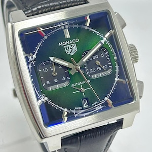 Puede incluir: Un reloj automático Monaco Tag Heuer de acero inoxidable con esfera cuadrada verde y azul. El reloj tiene una correa de cuero negro y múltiples subesferas. La esfera del reloj muestra el texto "MONACO TAG HEUER AUTOMATIC".