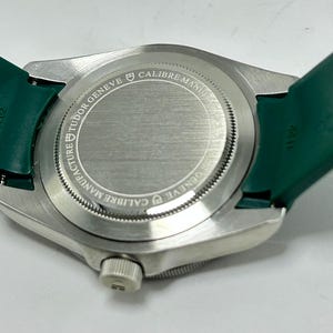 May include: Close-up of a silver wristwatch with a green band. The watch back is visible, displaying the text "TUDOR GENEVE CALIBRE MANUFACTURE". The green band is attached to the watch case.
