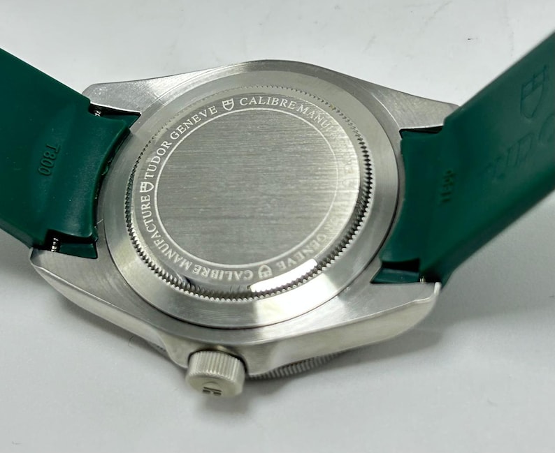 May include: Close-up of a silver wristwatch with a green band. The watch back is visible, displaying the text "TUDOR GENEVE CALIBRE MANUFACTURE". The green band is attached to the watch case.