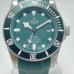 May include: A green and silver Tudor Pelagos watch with a green strap. The watch face is green with white markers and hands. The bezel is black with white numerals. The watch is water resistant to 600m.
