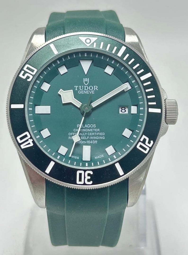 May include: A green and silver Tudor Pelagos watch with a green strap. The watch face is green with white markers and hands. The bezel is black with white numerals. The watch is water resistant to 600m.