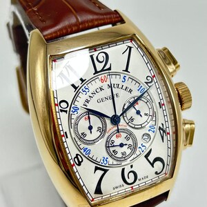 May include: A gold-toned tonneau-shaped wristwatch with a white guilloch&eacute; dial, black Arabic numerals, and three sub-dials. The watch features blue hands, a brown leather strap with a crocodile texture, and the text Franck Muller Geneve Swiss Made.