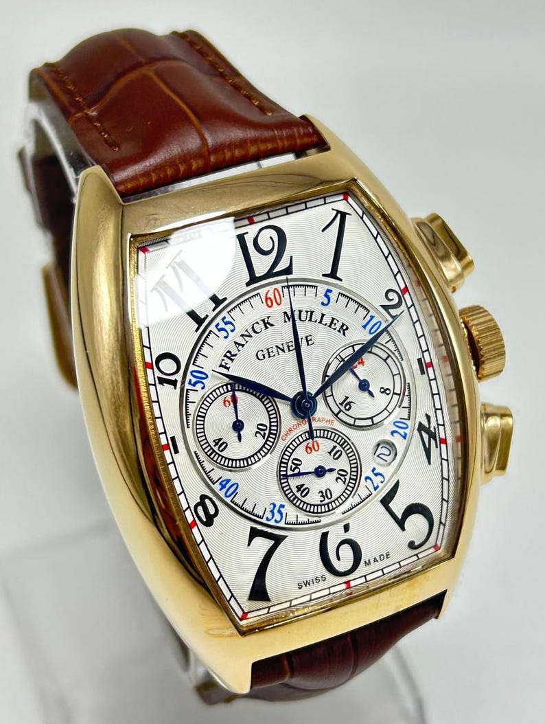 May include: A gold-toned tonneau-shaped wristwatch with a white guilloch&eacute; dial, black Arabic numerals, and three sub-dials. The watch features blue hands, a brown leather strap with a crocodile texture, and the text Franck Muller Geneve Swiss Made.