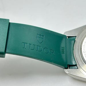 May include: Close-up of a green watch strap with the embossed word "TUDOR" and a shield logo. The watch's silver case is visible on the right. The strap is attached to the watch case with a metal clasp.