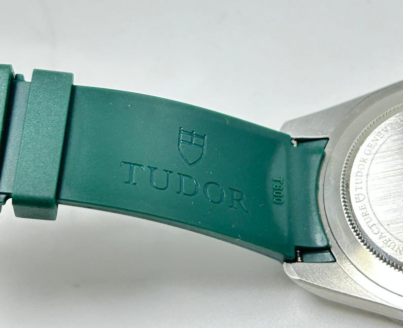 May include: Close-up of a green watch strap with the embossed word "TUDOR" and a shield logo. The watch's silver case is visible on the right. The strap is attached to the watch case with a metal clasp.