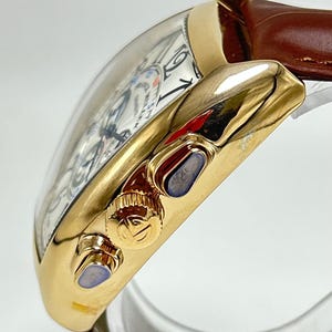 May include: A gold-toned wristwatch with a curved tonneau-shaped case and a brown leather strap. The side of the case features a textured crown with a logo and two pushers with blue accents. The white dial displays black numerals and text.
