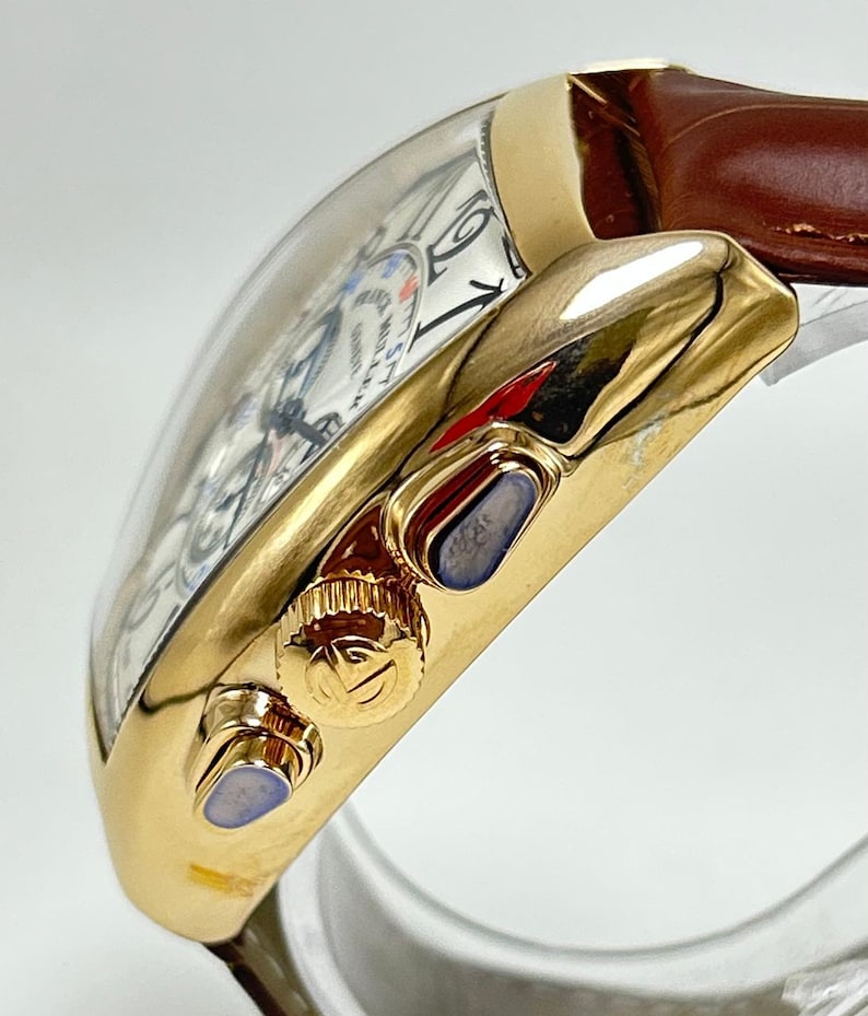 May include: A gold-toned wristwatch with a curved tonneau-shaped case and a brown leather strap. The side of the case features a textured crown with a logo and two pushers with blue accents. The white dial displays black numerals and text.