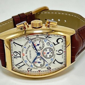 May include: A gold-toned tonneau-shaped chronograph wristwatch featuring a white dial with black Arabic numerals, three sub-dials, and blue hands. The watch includes a brown leather strap with a crocodile-embossed texture and a gold-toned buckle.