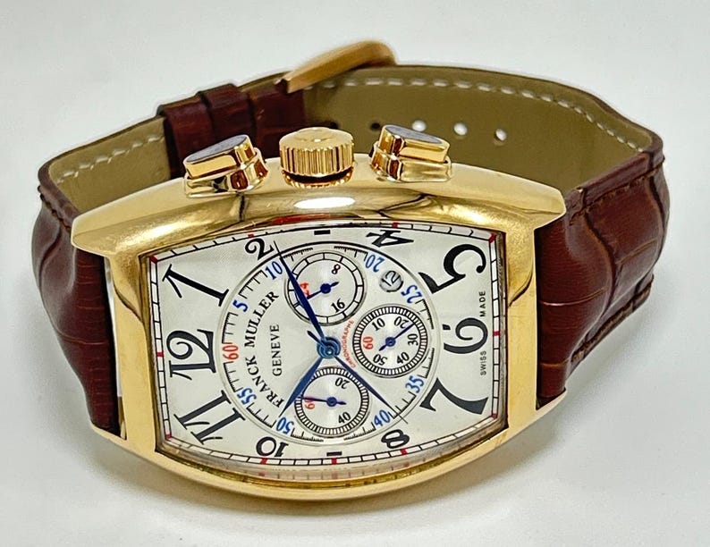 May include: A gold-toned tonneau-shaped chronograph wristwatch featuring a white dial with black Arabic numerals, three sub-dials, and blue hands. The watch includes a brown leather strap with a crocodile-embossed texture and a gold-toned buckle.