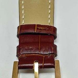 May include: A brown leather watch strap with a crocodile-embossed texture and white stitching. The strap features a gold-toned metal buckle with a small engraved logo at the base and two matching leather keepers to secure the end of the band.