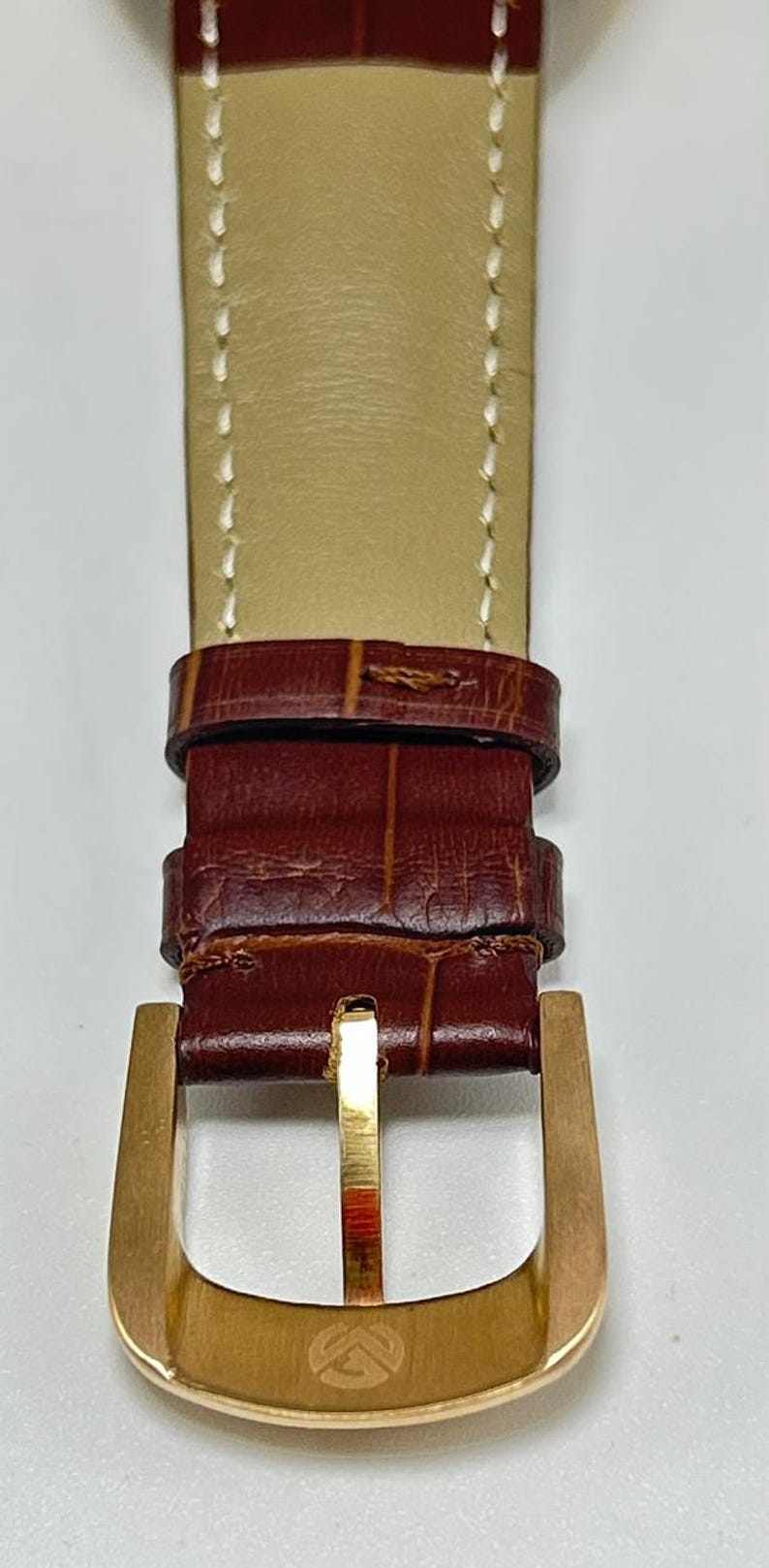 May include: A brown leather watch strap with a crocodile-embossed texture and white stitching. The strap features a gold-toned metal buckle with a small engraved logo at the base and two matching leather keepers to secure the end of the band.