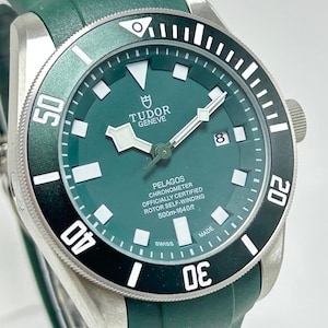 May include: A close-up of a Tudor Pelagos watch with a green dial and bezel. The watch has a stainless steel case and a green rubber strap. The dial features white hour markers and hands, with the date at the 3 o'clock position. The watch is water-resistant to 1640 feet.