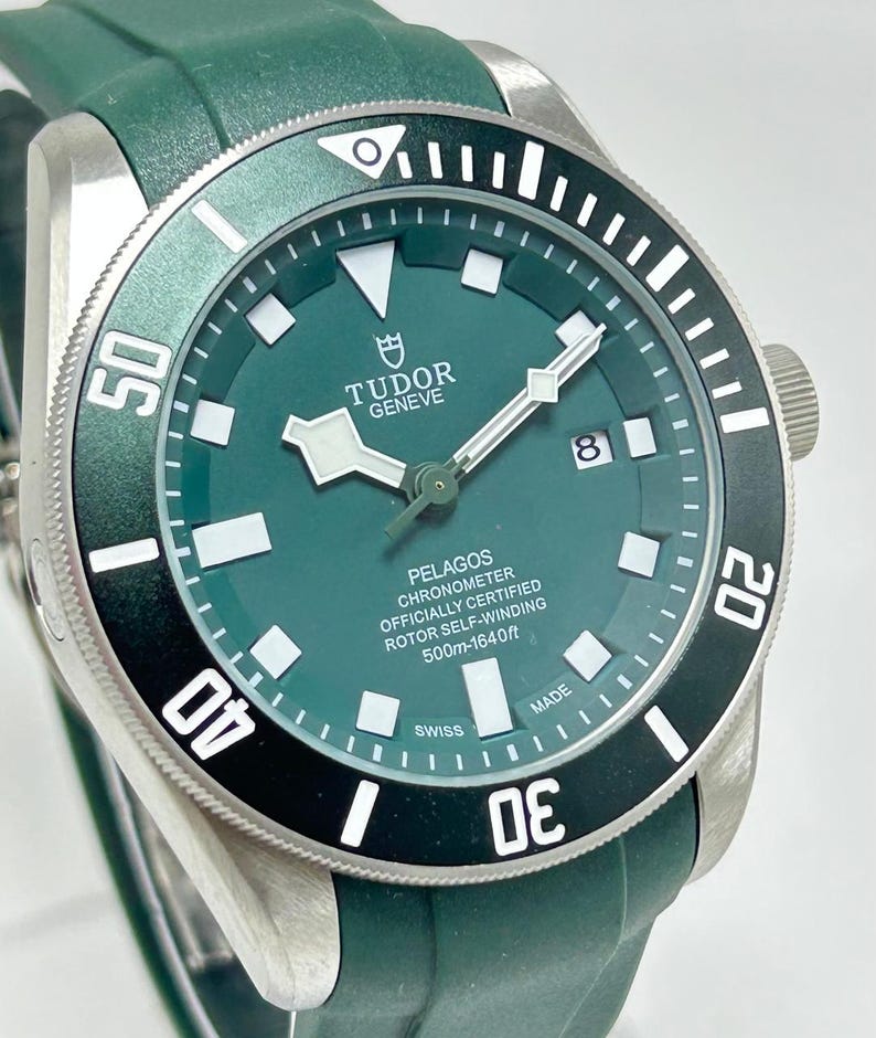 May include: A close-up of a Tudor Pelagos watch with a green dial and bezel. The watch has a stainless steel case and a green rubber strap. The dial features white hour markers and hands, with the date at the 3 o'clock position. The watch is water-resistant to 1640 feet.