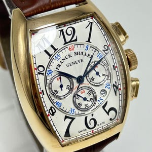 Könnte beinhalten: Eine goldfarbene tonnenförmige Chronographenuhr mit weißem Guilloché-Zifferblatt, schwarzen arabischen Ziffern und drei Hilfszifferblättern. Die Uhr trägt den Schriftzug Franck Muller Geneve und ein braunes Lederarmband mit Krokodilprägung.