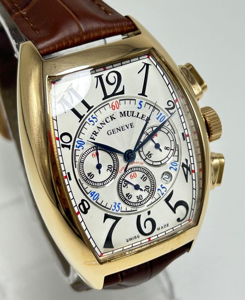 May include: A gold-toned tonneau-shaped chronograph watch with a white guilloch&eacute; dial, black Arabic numerals, and three sub-dials. The watch features the text Franck Muller Geneve and a brown leather strap with a visible crocodile-embossed texture.