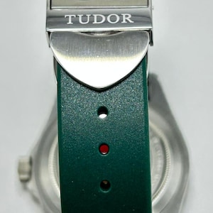 May include: Close-up of a green watch strap with a silver clasp. The clasp is engraved with the word "TUDOR". The watch strap is made of a textured material. The watch face is visible in the background.