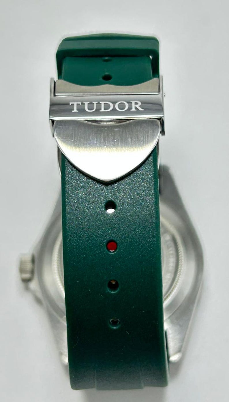 May include: Close-up of a green watch strap with a silver clasp. The clasp is engraved with the word "TUDOR". The watch strap is made of a textured material. The watch face is visible in the background.