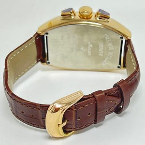 May include: A gold-toned tonneau-shaped wristwatch with a brown embossed leather strap and a gold-toned buckle. The watch case features two side pushers and a crown. The back of the case is engraved with the text Franck Muller Geneve.