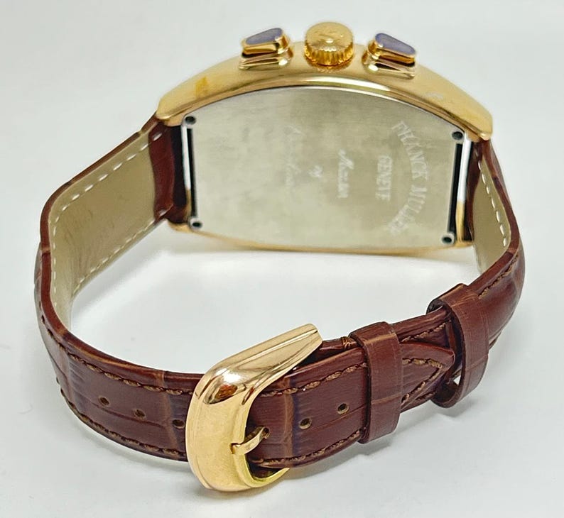 May include: A gold-toned tonneau-shaped wristwatch with a brown embossed leather strap and a gold-toned buckle. The watch case features two side pushers and a crown. The back of the case is engraved with the text Franck Muller Geneve.