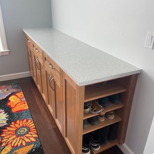 May include: A wooden cabinet with a white countertop and open shelves below. The shelves are filled with shoes. The cabinet is in a hallway with a patterned rug on the floor.