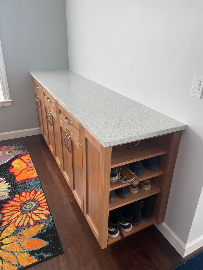 May include: A wooden cabinet with a white countertop and open shelves below. The shelves are filled with shoes. The cabinet is in a hallway with a patterned rug on the floor.