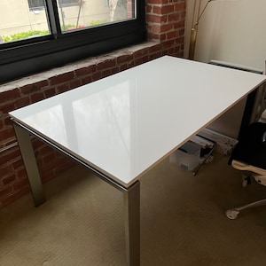May include: A white rectangular table with a silver metal frame. The table is sitting on a beige carpet. The table is in a room with a brick wall and a window.