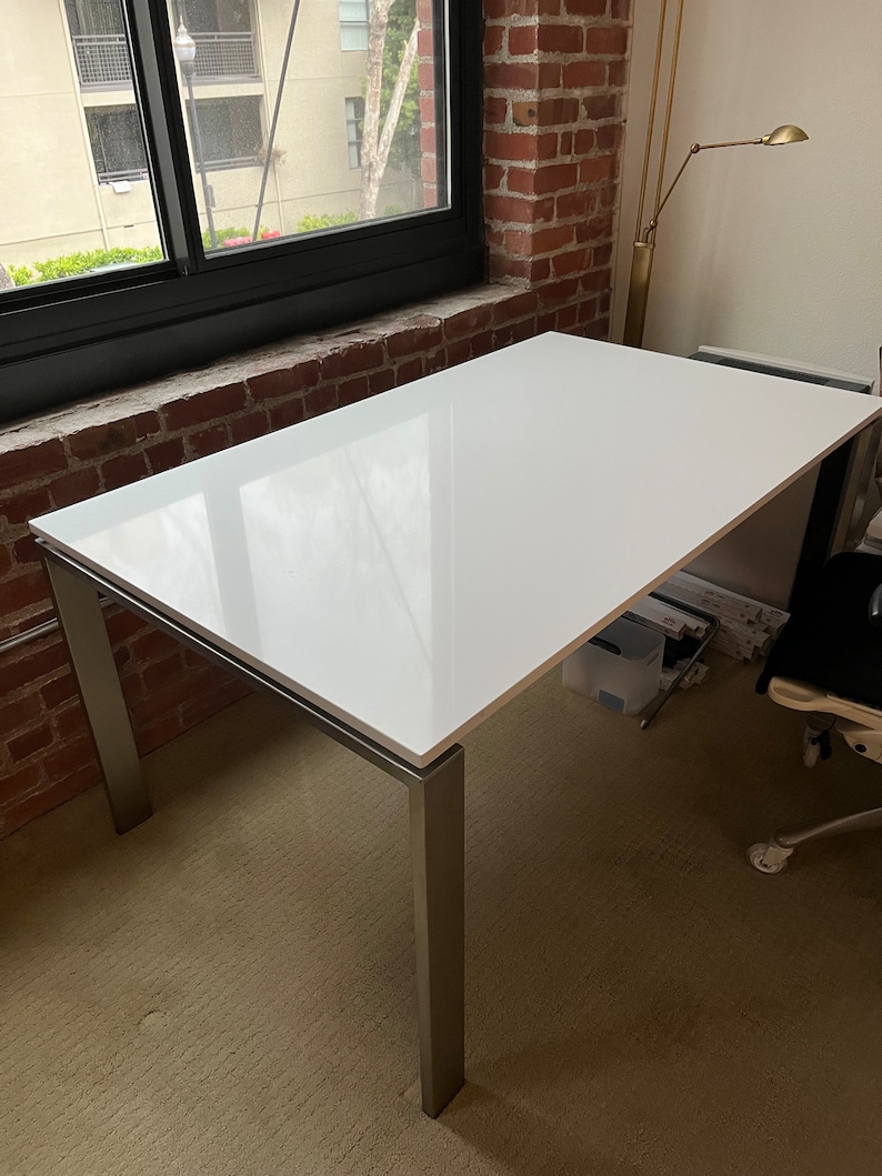 May include: A white rectangular table with a silver metal frame. The table is sitting on a beige carpet. The table is in a room with a brick wall and a window.