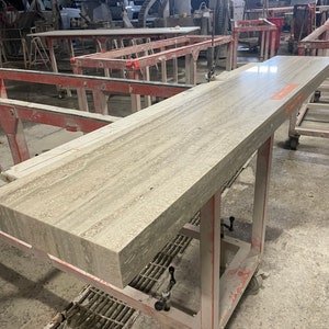 May include: A large, light gray countertop slab with a wood-like grain pattern sits on a red metal rolling cart with black wheels. The slab is in a workshop setting.