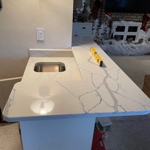 May include: A white countertop with a grey veined pattern, a stainless steel sink, and a yellow spirit level. The countertop is part of a bar or kitchen island.
