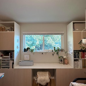 May include: A white desk with a light wood laminate top and a white printer on the surface. The desk is flanked by two white cabinets with light wood laminate doors. The cabinets have vertical grooves. A white window with a view of trees is above the desk. A white board with the text "Make Up Your Mind Think Work Hard Hustle Own Salty Worry Less Be Happy" is on the right side of the desk.