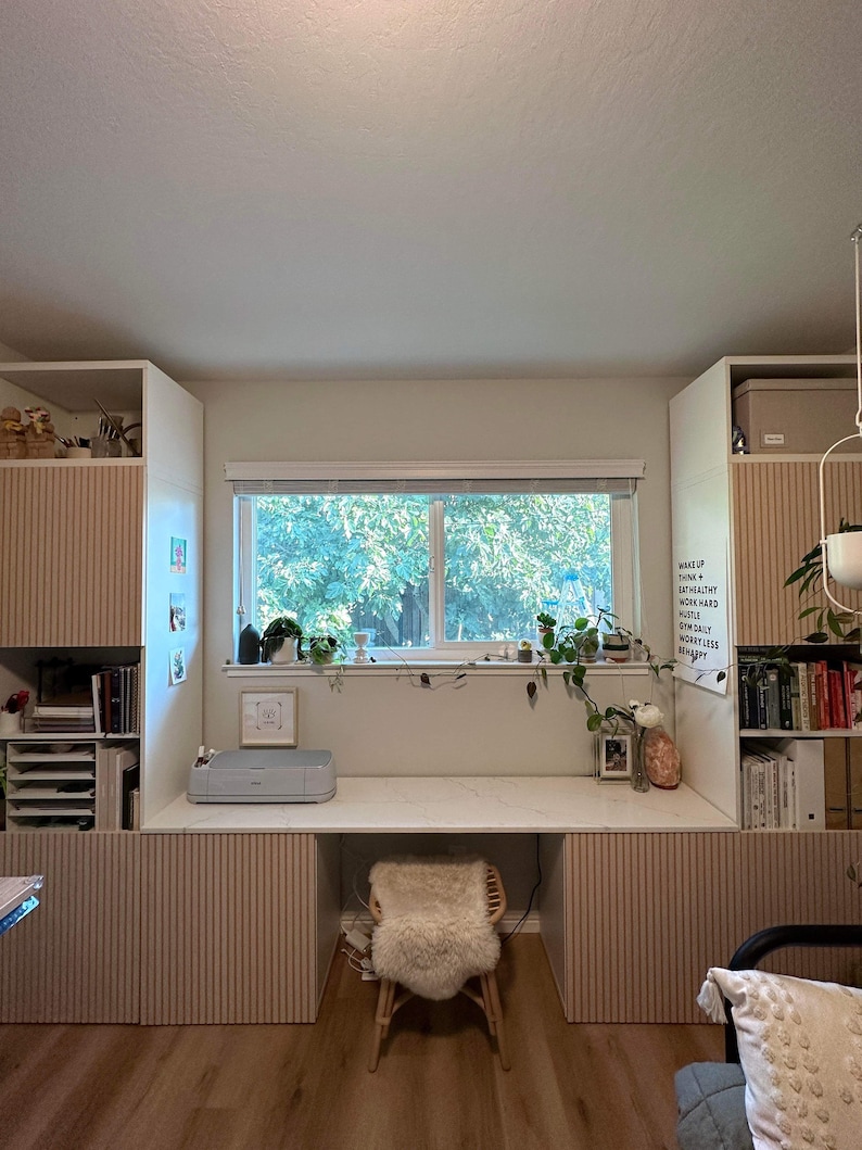 May include: A white desk with a light wood laminate top and a white printer on the surface. The desk is flanked by two white cabinets with light wood laminate doors. The cabinets have vertical grooves. A white window with a view of trees is above the desk. A white board with the text "Make Up Your Mind Think Work Hard Hustle Own Salty Worry Less Be Happy" is on the right side of the desk.