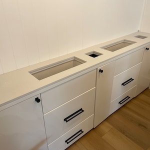 May include: White kitchen cabinets with a white worktop and black hardware. The cabinets have three drawers and a door. The worktop has three cutouts for sinks or other fixtures.