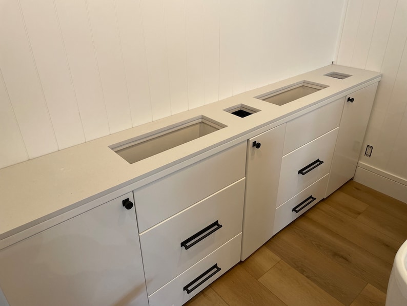 May include: White kitchen cabinets with a white worktop and black hardware. The cabinets have three drawers and a door. The worktop has three cutouts for sinks or other fixtures.